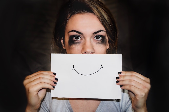 A woman with smeared eye makeup holds a piece of paper with a smile on it in front of her face